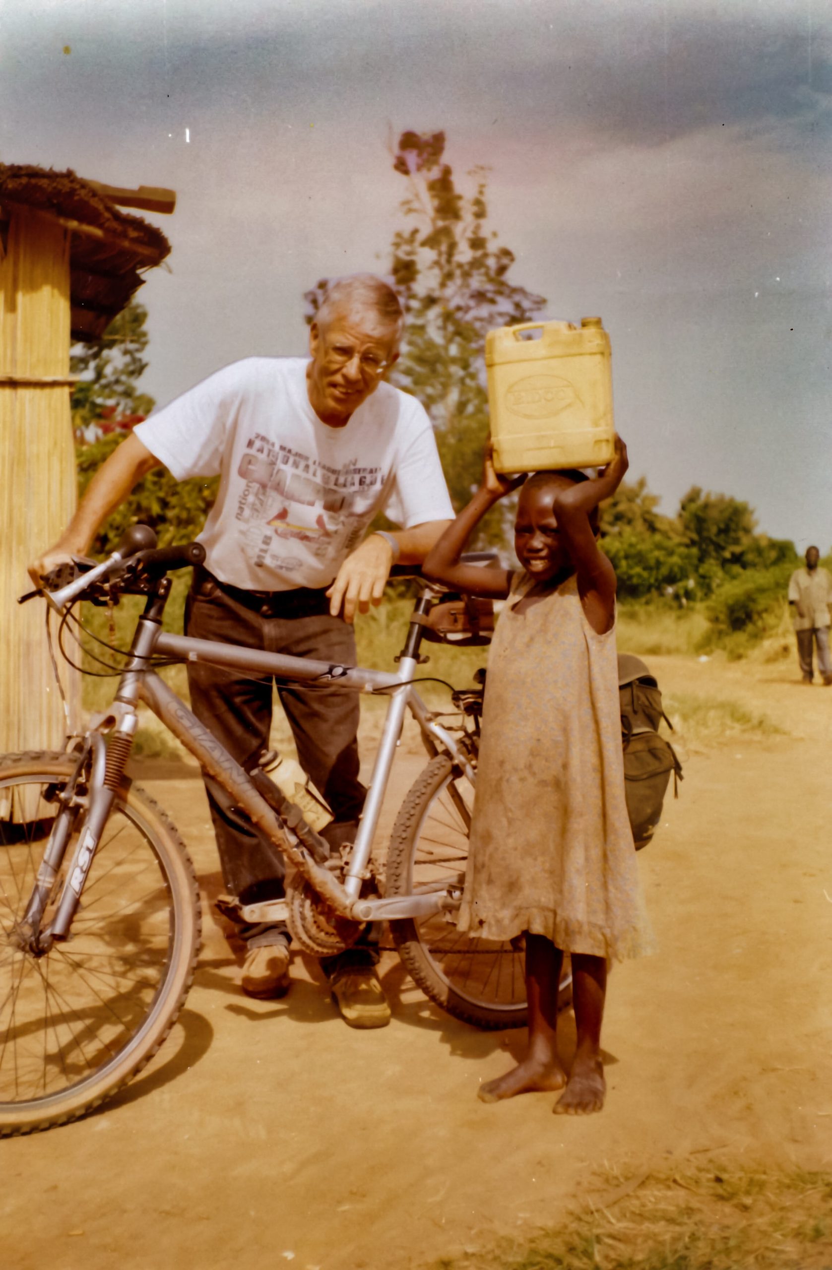 Fr David Baltz leans on his bicycle. A young African girls wearing a tattered dress holds a jerry can of water on her head. she is standing next to the bike.