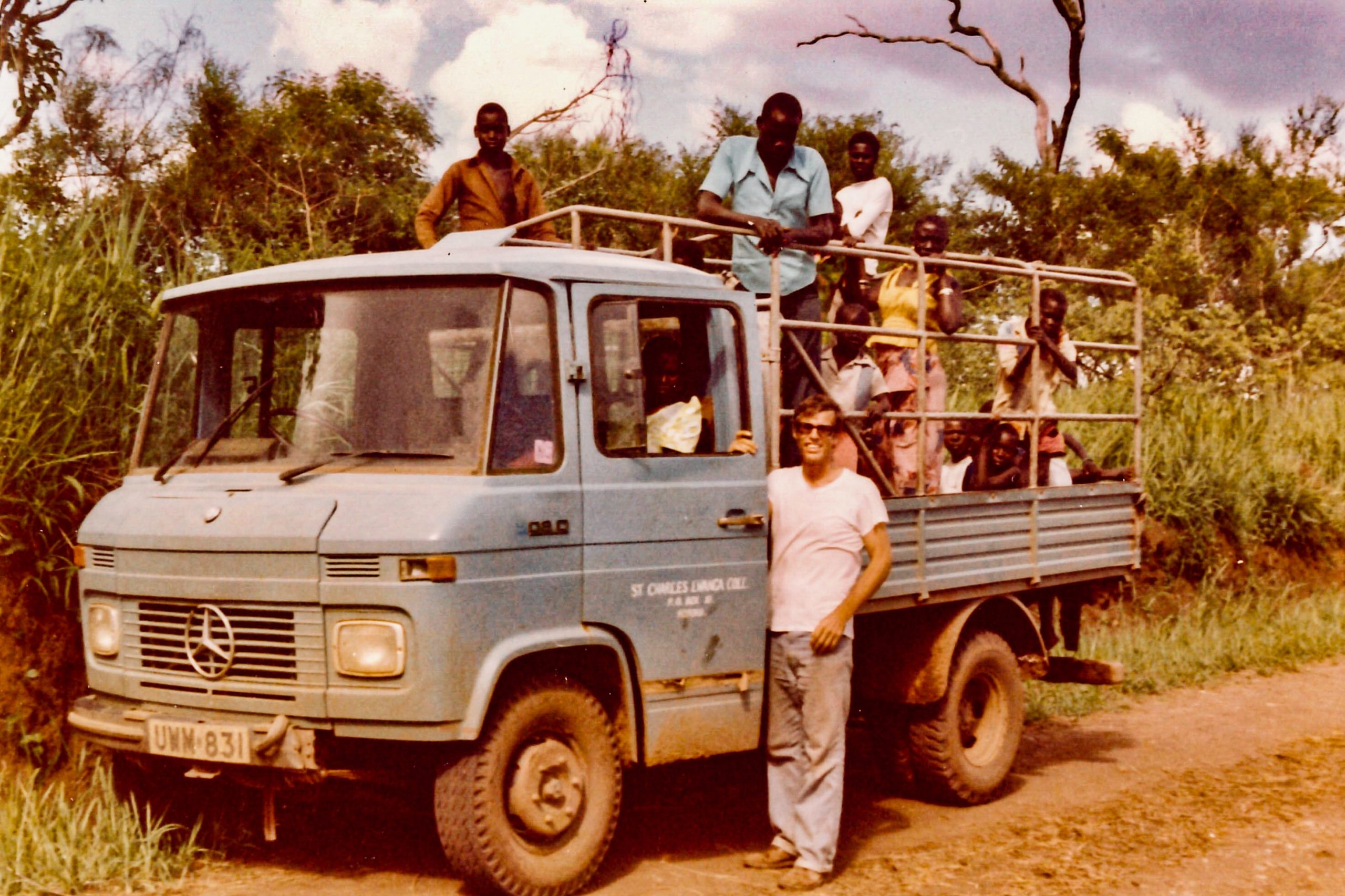 Fr David Baltz stands next to a mercedes lorry in a photo from the 1970s the lorry is open air in the back and several Africans are riding. the side of the lorry says St Charles Lwanga