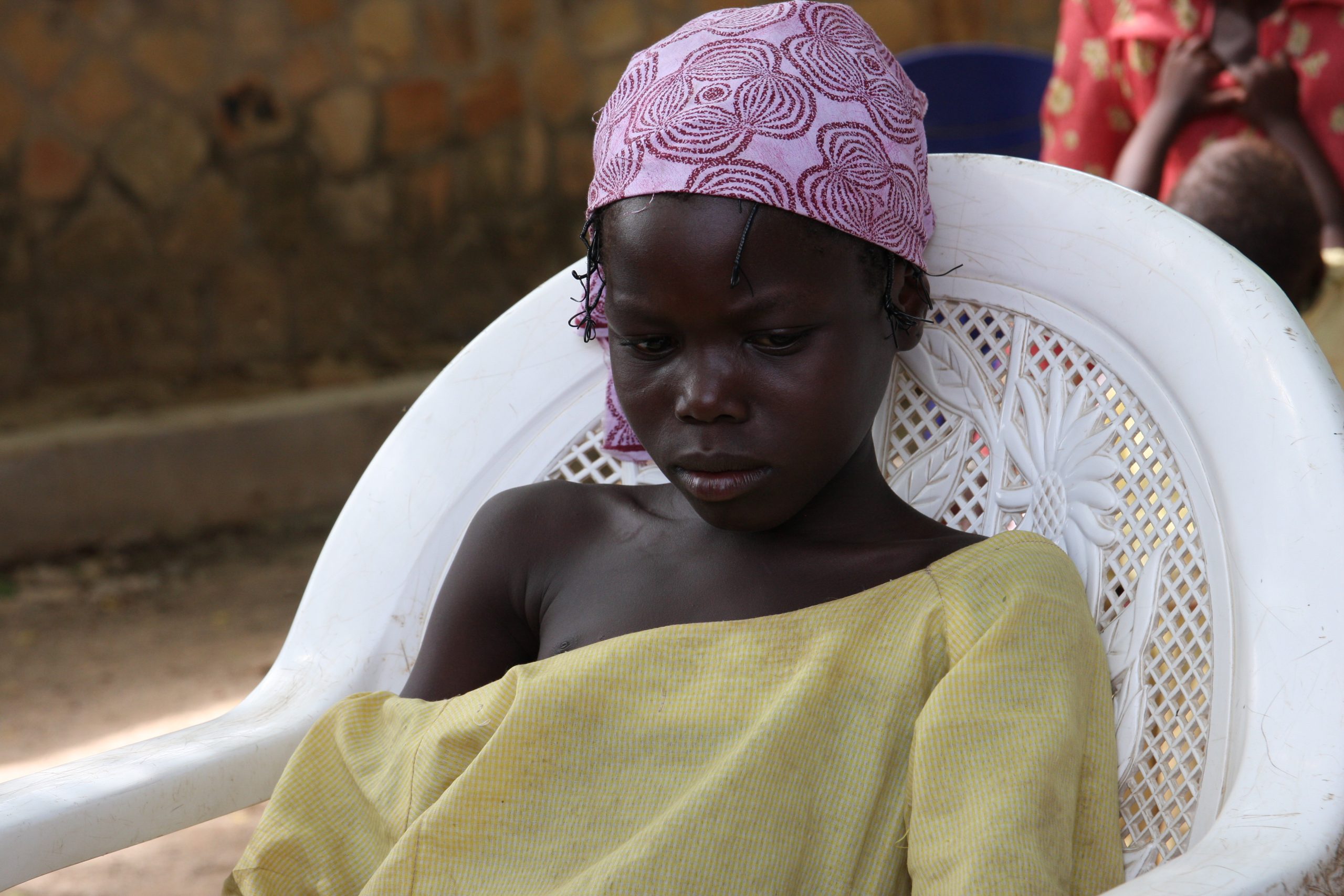 A young Nigerian Girl wearing an oversized dirty yellow dress, a pink and purple scarf on her head, sits in a white plastic chair