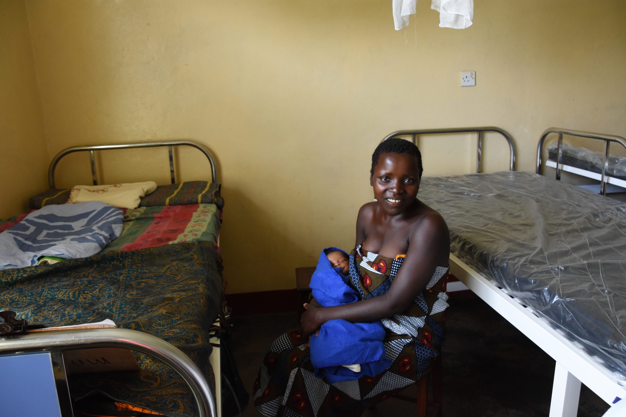 A Ugandan mother holds her newborn baby wrapped in a blue blanket She sits next to a new maternity bed recently donated to Matany hospital