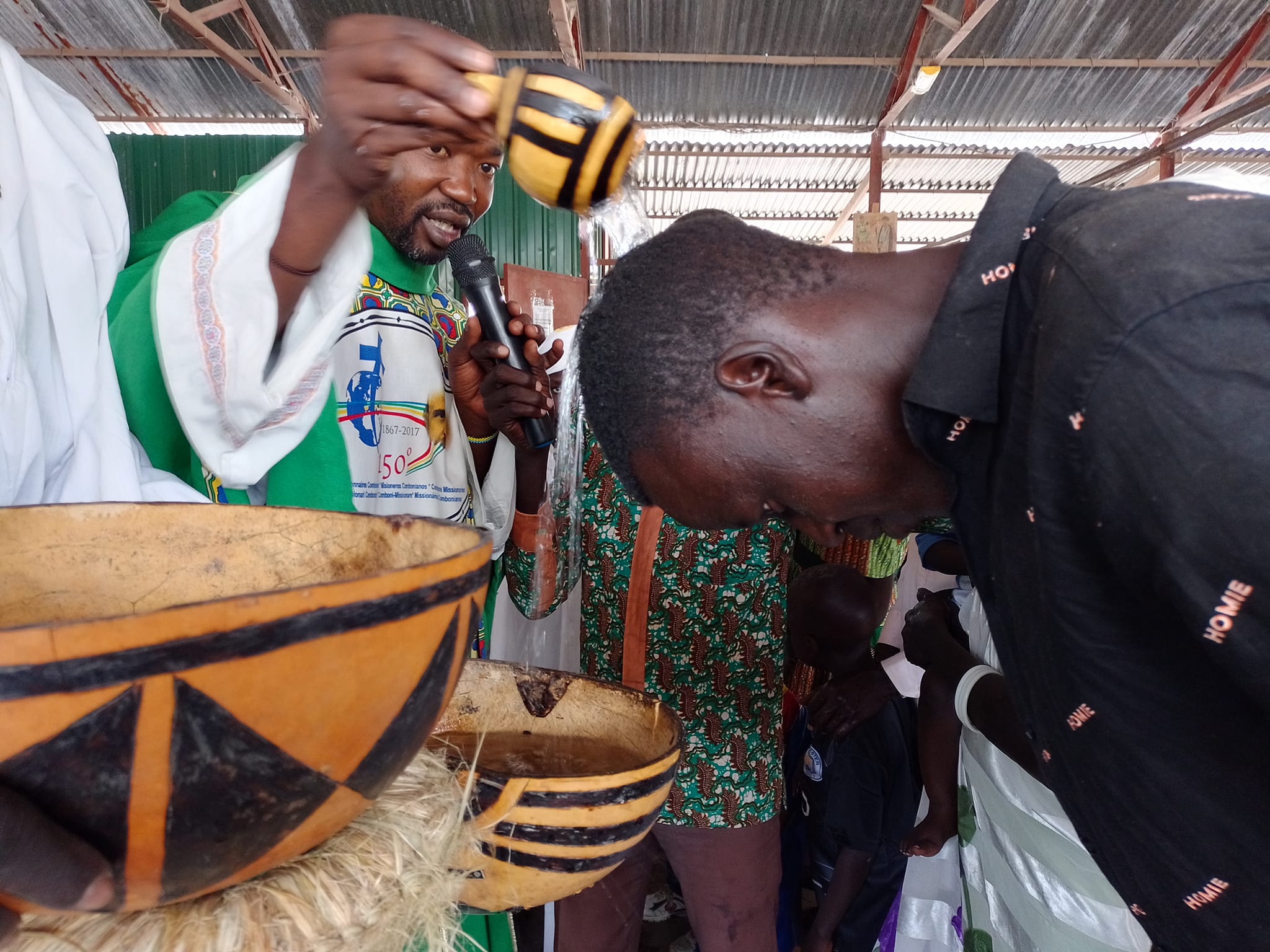Fr Alfred pours water over a mans head for baptism. The baptismal cup is made from a gourd. the bowl beneath the young man is wooden with traditional African carvings