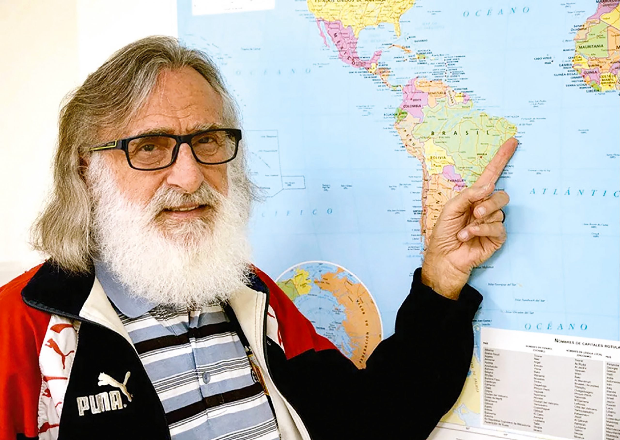 Fr Carlos Basacarn pointing to Africa on a map. He has a long, white beard and is wearing dark rimmed glasses.