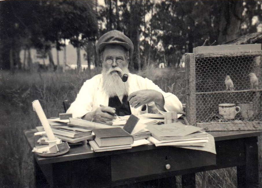 Fr Zorn, also called Baba Zulu, smokes a pipe at his desk outside in the 1920s. A bird cage is behind him.
